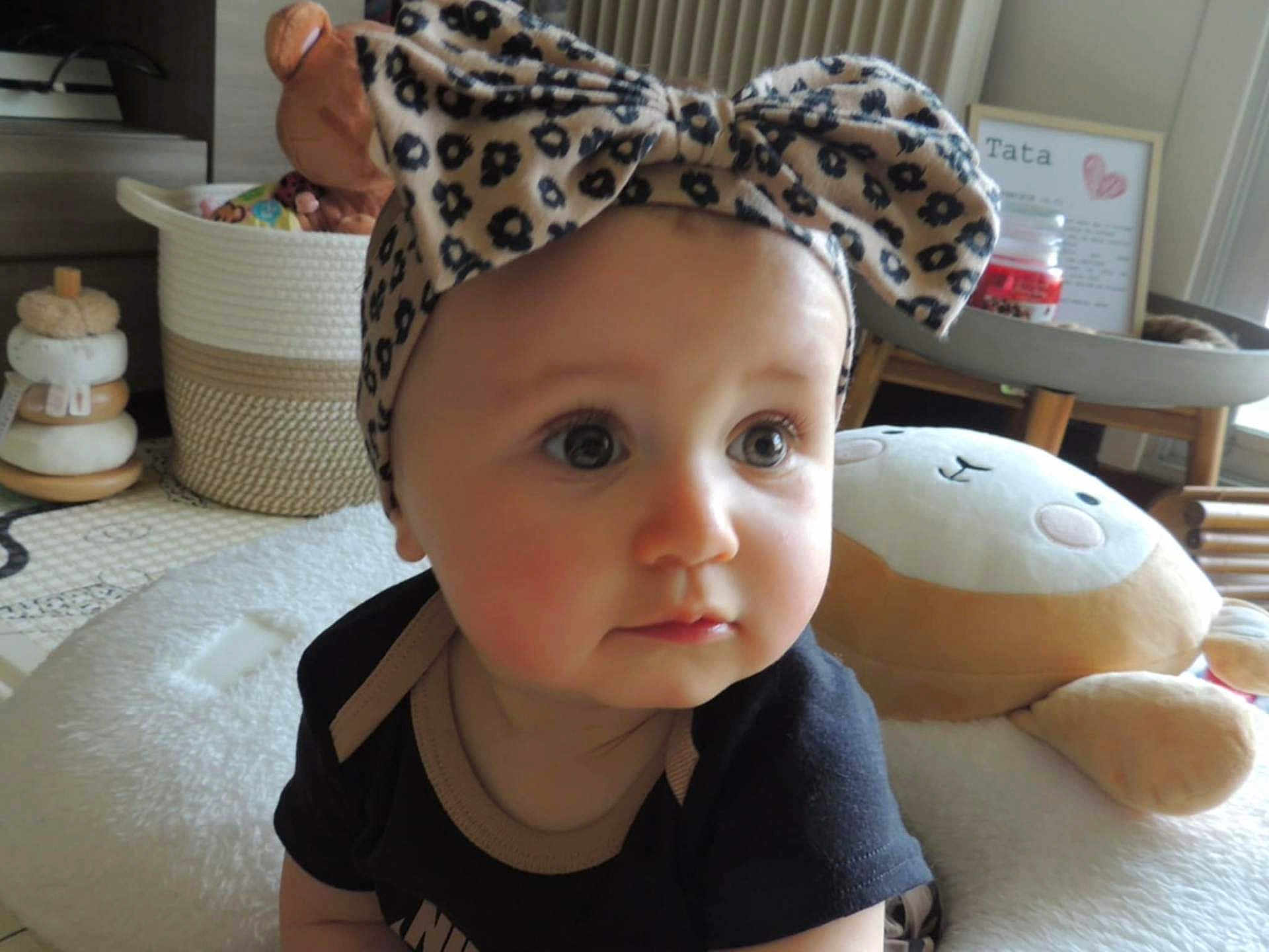 Alya a rejoint le concours — aidez-le/la à gagner de superbes lots ! baby, headband, bow, face, child, plush_toy, cushion, indoor, soft_light, curious_expression, home_decor, basket, toy, infant, black_clothing, closeup, seated, animal_plush, floor_mat, room