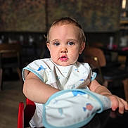 Anastasia is registered to the contest to win money with this photo: arm, baby, bib, blurred_background, casual, child, clothing, curious, drool, expression, face, high_chair, indoor, light, person, portrait, seat, table, toddler, young_child