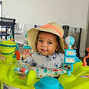 Emilia joined the competition — help win amazing prizes! baby, child, hat, activity_center, toys, indoor, smiling, face, person, chair, clock, table, colorful, playful, infant, happy, cute, furniture, room, portrait