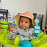 baby, child, hat, activity_center, toys, indoor, smiling, face, person, chair, clock, table, colorful, playful, infant, happy, cute, furniture, room, portrait