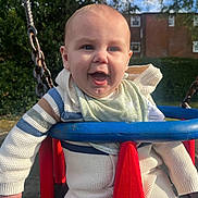 Ruben is registered to the contest to win money with this photo: baby, child, swing, playground, outdoor, smiling, happy, clothing, sweater, bib, sunlight, daytime, chains, red, blue, greenery, residential_building, face, person, fun