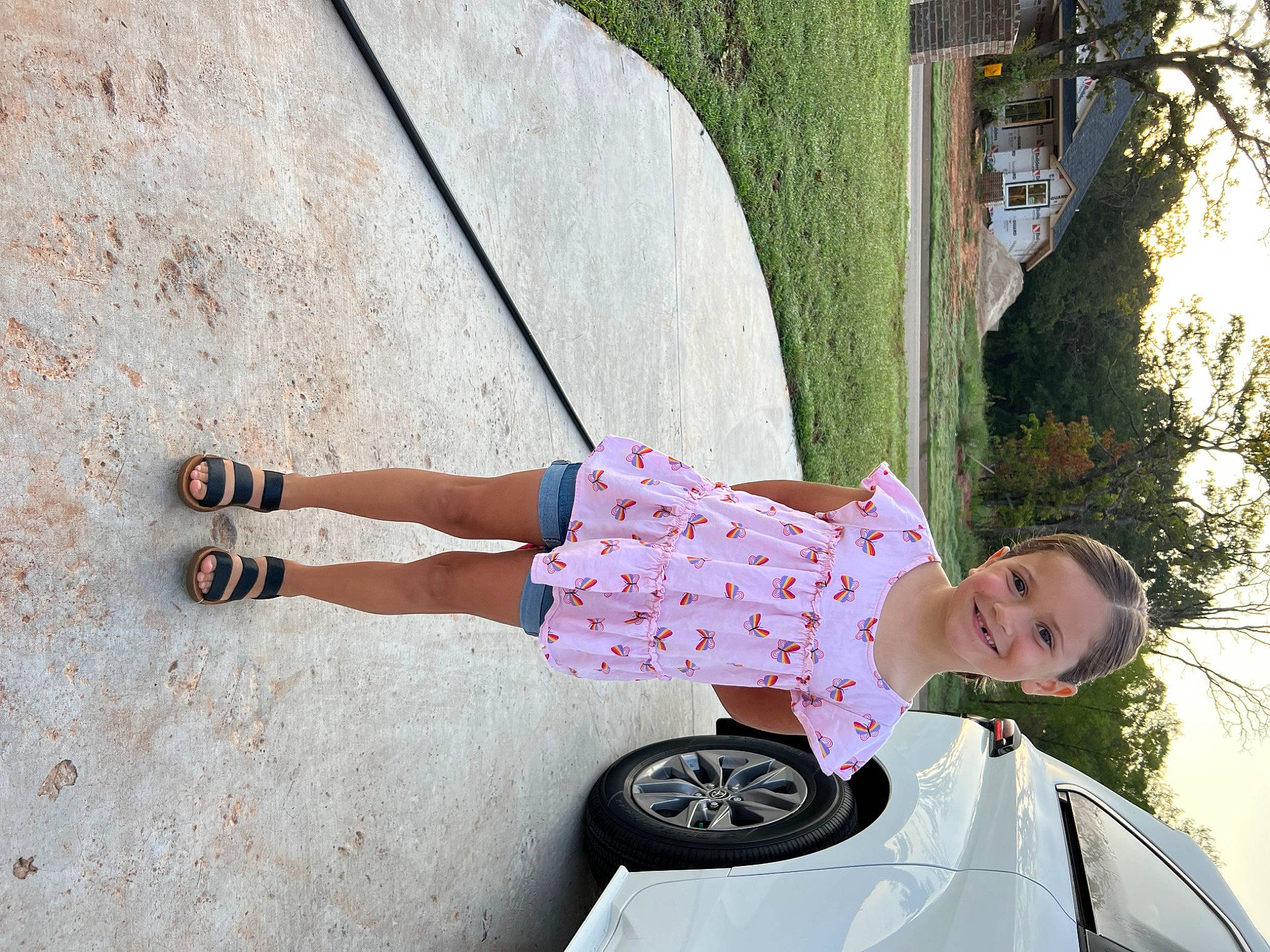 Tatum is registered to the contest to win money with this photo: automotive_exterior, automotive_tire, car, dress, fender, footwear, fun, hood, joy, motor_vehicle, person, pink, plant, red, summer, tints_and_shades, tire, toddler, tree, vehicle