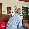 back_view, casual_clothing, cozy, dog, embrace, friendly, furniture, gray_sweater, home, indoor, man, pet, plant, red_couch, relaxing, table, trunk, wall_art, window, wooden_paneling
