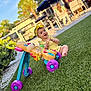 baby, child, toy, walker, grass, outdoor, sunlight, yellow_clothing, headband, purple_wheels, blue_handle, smiling, playful, patio, chairs, umbrellas, greenery, happy, person, daytime