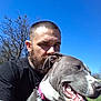 man, dog, beard, tattoo, outdoor, blue_sky, tree, collar, pet, happy, tongue_out, grass, sunlight, casual_clothing, animal, nature, portrait, smile, friendship, daytime