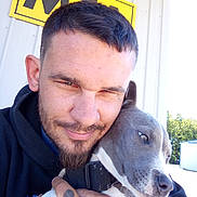 Freya is registered to the contest to win money with this photo: man, dog, pet, outdoor, building, tattoo, smile, short_hair, beard, collar, close_up, hug, sunlight, casual_clothing, fence, expression, face, friendship, animal, human