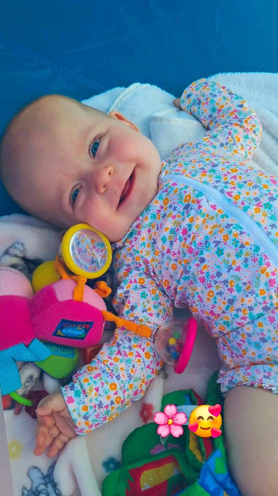 Tyana participe au concours pour gagner de l'argent avec cette photo : baby, infant, smile, floral_onesie, toy, blanket, colorful, playful, happy, cute, face, child, lying_down, soft_texture, indoors, joyful, cheerful, closeup, person, emotion