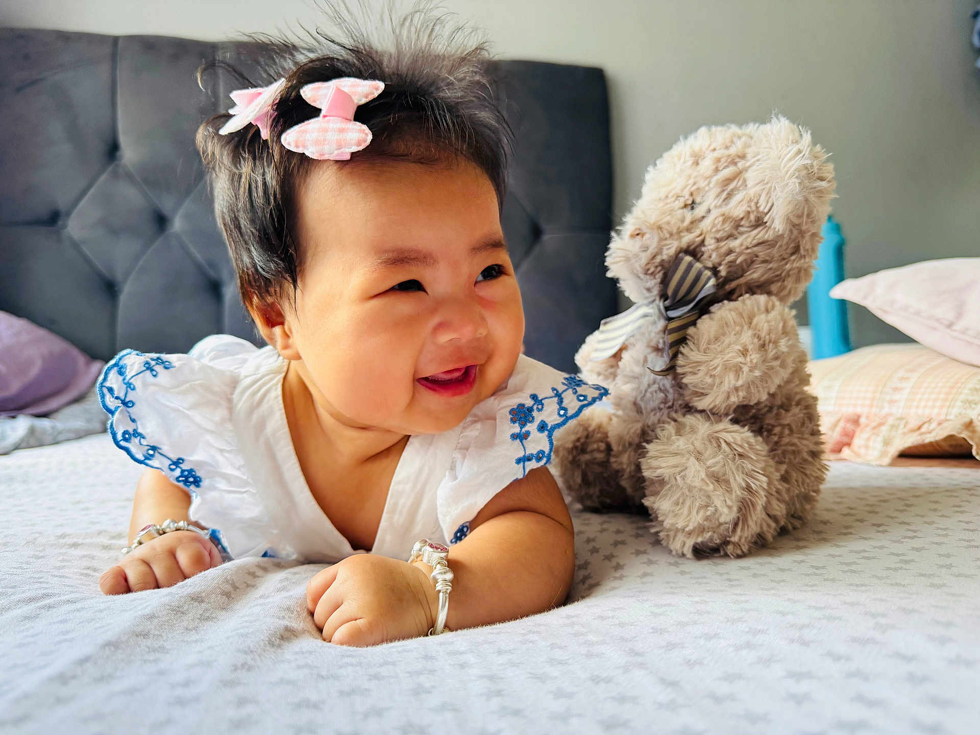 Yulin is registered to the contest to win money with this photo: baby, bed, bracelet, child, cute, embroidered, furniture, hair_bows, happy, headboard, indoor, infant, person, pillow, playful, plush_toy, smiling, soft_toy, teddy_bear, white_dress