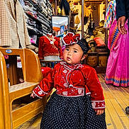 Yulin Fung Tumbapo is registered to the contest to win money with this photo: child, toddler, traditional_clothing, red_dress, black_skirt, headband, shop_interior, fabric, baskets, wooden_floor, mannequin, chair, person, colorful, textile, earrings, portrait, curious, indoor, decor