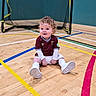 toddler, child, soccer_uniform, sports_court, goalpost, indoor, curly_hair, maroon_shirt, white_shorts, white_socks, white_shoes, floor_lines, smiling, sitting, young_child, sports_equipment, playful, athletic, cute, portrait