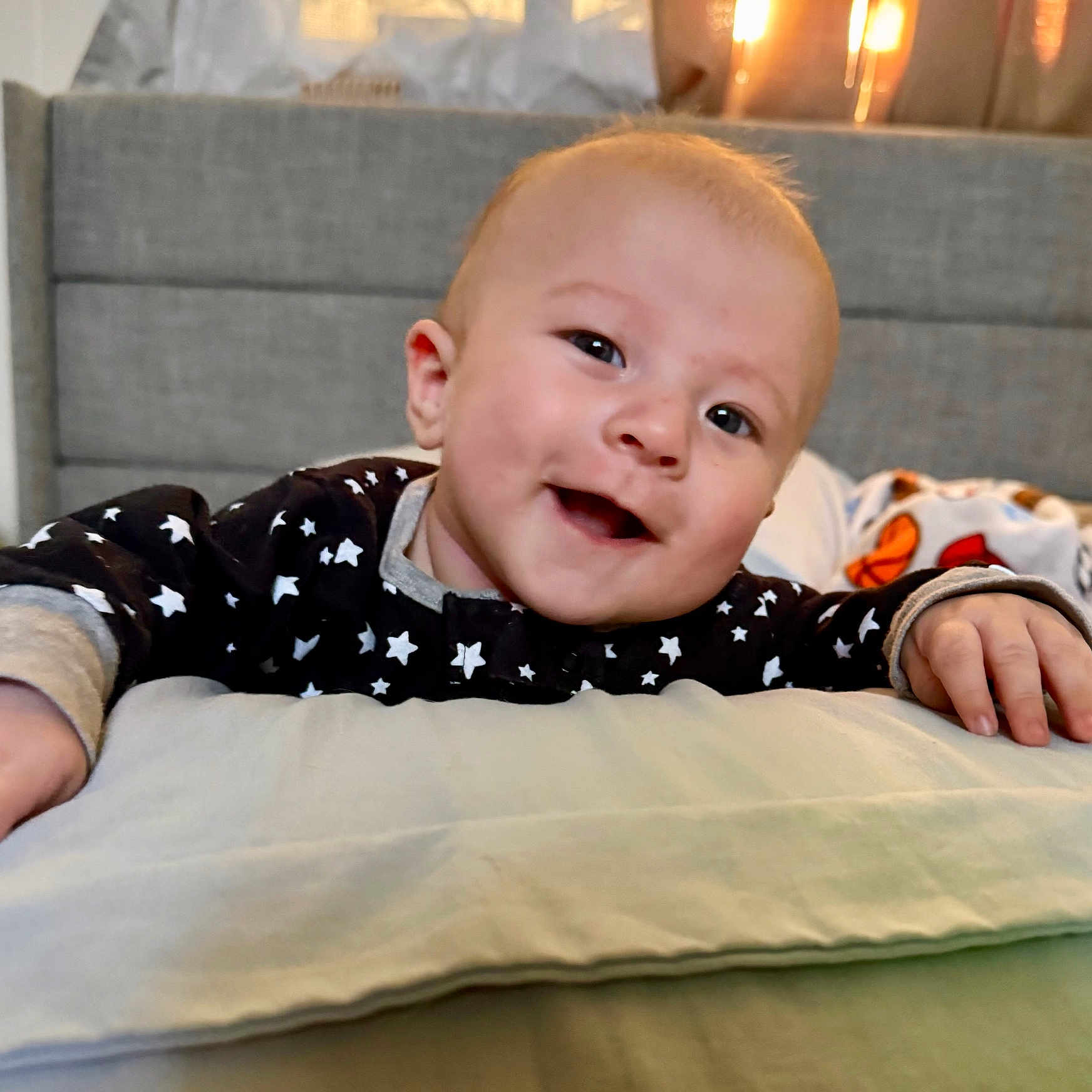 Theo joined the competition — help win amazing prizes! baby, smile, face, infant, pillow, hand, star_pattern, clothing, indoors, bedroom, blanket, cute, happy, child, head, young, person, warm_light, furniture, casual