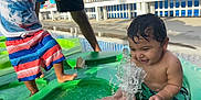 Carmelo is registered to the contest to win money with this photo: toddler, child, water, splash, pool, outdoor, summer, play, happy, person, green, shorts, swimwear, fence, sunlight, adult, hand_holding, wet_hair, foot, smile
