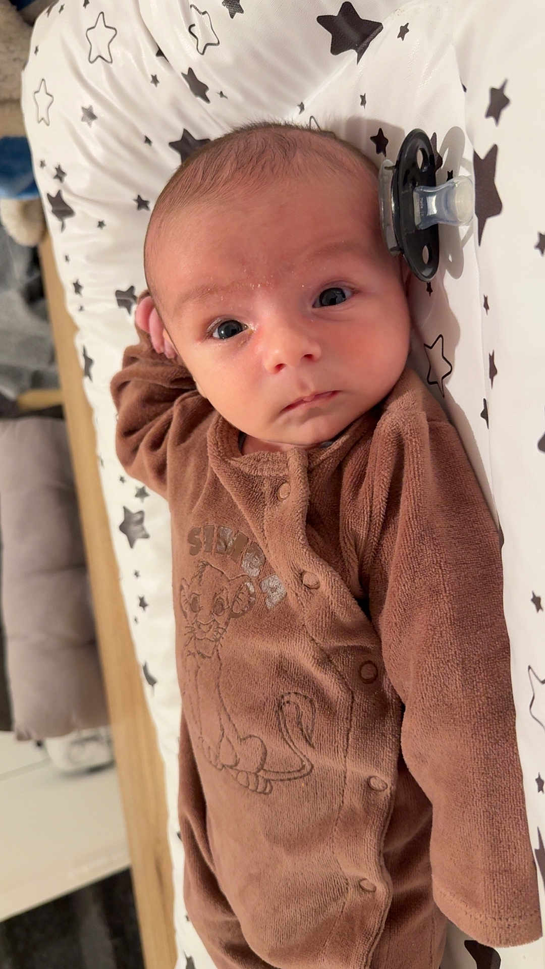 Liam participe au concours pour gagner de l'argent avec cette photo : baby, infant, onesie, brown_clothing, cartoon_lion, pacifier, star_pattern, cushion, lying_down, wide_eyes, face, hand, cute, indoor, wooden_surface, soft_texture, child, newborn, portrait, looking