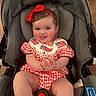 baby, child, car_seat, red_bow, tongue_out, smiling, cheeks, clothing, red_white_checkered, american_flag, headband, indoor, seat_belt, infant, cute, playful, person, portrait, happy, baby_girl