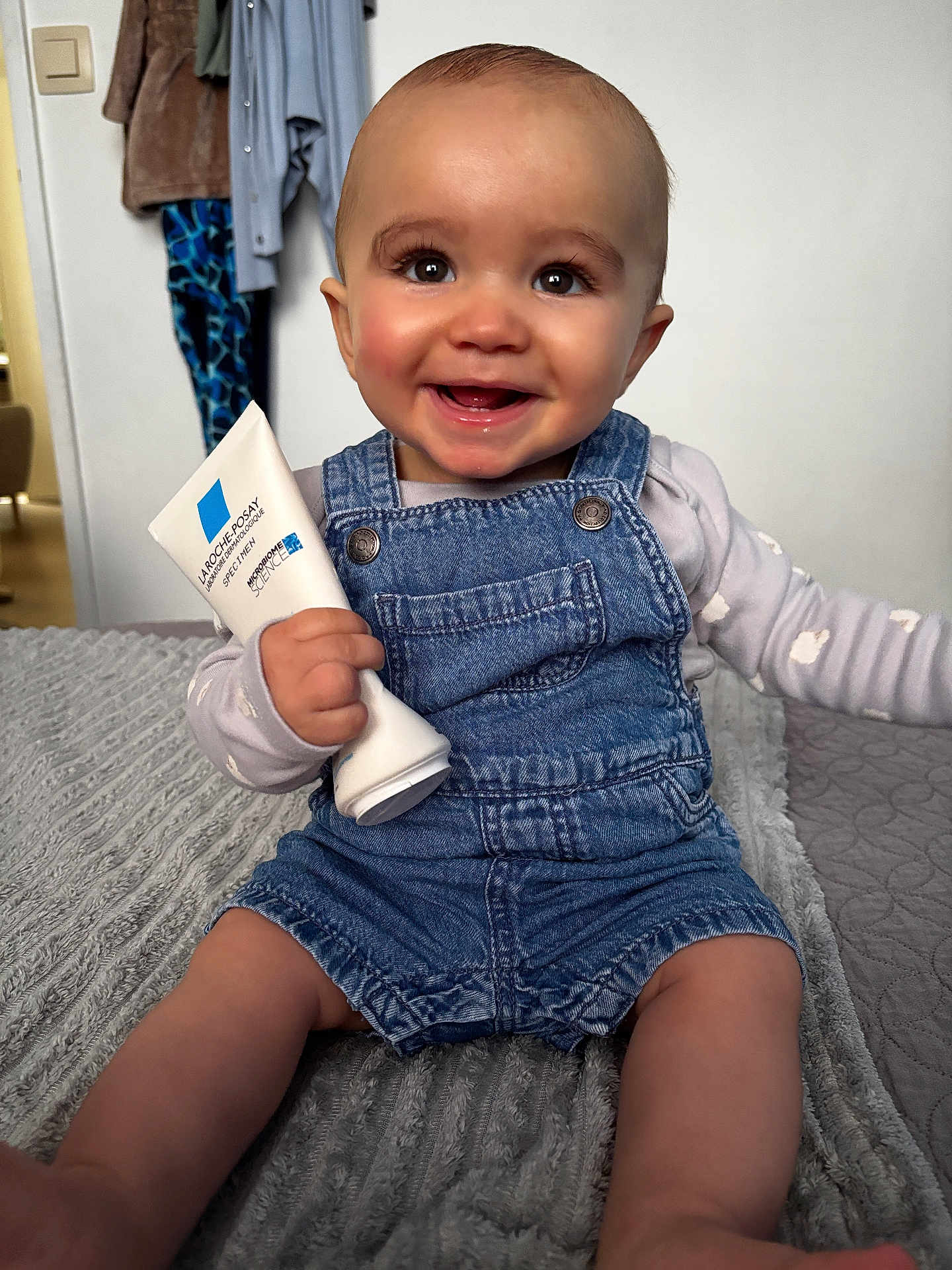 Elyo participe au concours pour gagner de l'argent avec cette photo : baby, smile, denim_overalls, cream_tube, indoor, blanket, clothing, happy, face, person, child, skin_care, furniture, room, head, sitting, cute, young_child, holding, casual