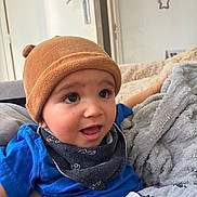 Elyo a rejoint le concours — aidez-le/la à gagner de superbes lots ! baby, child, hat, blue_shirt, scarf, blanket, couch, indoor, face, expression, curious, seated, cozy, soft_texture, comfort, person, young_child, clothing, headwear, home