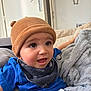 baby, child, hat, blue_shirt, scarf, blanket, couch, indoor, face, expression, curious, seated, cozy, soft_texture, comfort, person, young_child, clothing, headwear, home