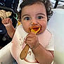 baby, child, high_chair, bib, spoon, food, messy, cute, face, happy, indoor, clothing, white_bow, curly_hair, person, feeding, close_up, infant, smiling, table