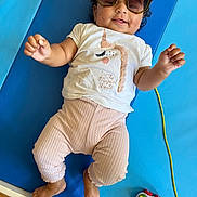 Emma a rejoint le concours — aidez-le/la à gagner de superbes lots ! baby, child, sunglasses, unicorn, shirt, pink_pants, blue_mat, toy_car, smiling, lying_down, indoor, cute, happy, person, feet, hand, curly_hair, playtime, casual_clothing, infant