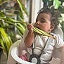 baby, child, high_chair, leaf, plant, indoor, curly_hair, chewing, white_chair, pink_shorts, patterned_shirt, straps, harness, natural_light, candid, exploration, cute, toddler, home, greenery