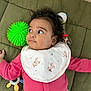 baby, infant, child, pink_clothing, bib, unicorn_pattern, green_toy, rattle, blanket, quilt, lying_down, curious_expression, indoors, playtime, cute, soft_lighting, portrait, face, hand, floor
