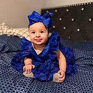 Mia joined the competition — help win amazing prizes! baby, child, blue_dress, headband, bow, bedspread, pattern, happy, smiling, tongue_out, sitting, indoor, cute, infant, hands, feet, soft_focus, bed, fabric, portrait