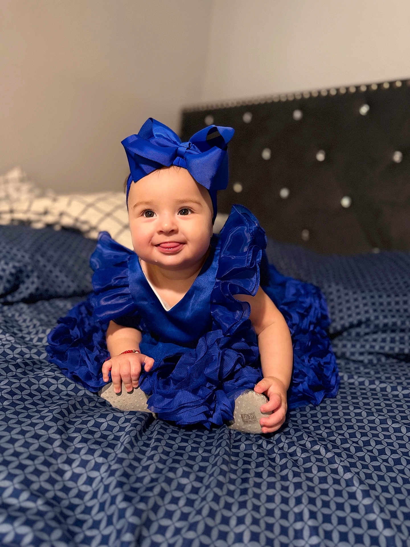 Mia joined the competition — help win amazing prizes! baby, child, blue_dress, headband, bow, bedspread, pattern, happy, smiling, tongue_out, sitting, indoor, cute, infant, hands, feet, soft_focus, bed, fabric, portrait