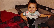 Raymason is registered to the contest to win money with this photo: toddler, child, playpen, bed, blanket, quilt, indoor, smiling, curly_hair, person, clothing, furniture, happy, baby, face, head, hands, fabric, blanket_pattern, cozy