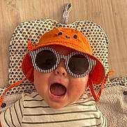 Andréas participe au concours pour gagner de l'argent avec cette photo : baby, child, orange_hat, sunglasses, striped_shirt, smiling, lying_down, hand, camera, patterned_mat, floor, cute, happy, infant, accessory, headwear, person, indoor, portrait, baby_clothes