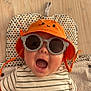 baby, child, orange_hat, sunglasses, striped_shirt, smiling, lying_down, hand, camera, patterned_mat, floor, cute, happy, infant, accessory, headwear, person, indoor, portrait, baby_clothes