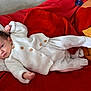 baby, infant, white_sweater, white_pants, red_blanket, pillow, lying_down, indoors, cozy, soft_fabric, cute, small_hands, face, head, newborn, child, resting, blanket, warm, comfort
