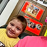 child, smiling, yellow_shirt, indoor, wood_panel_wall, photo_collage, happy, person, portrait, candid, home, casual, face, young_child, seated, colorful, wall_decor, family_photos, interior, people