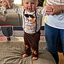 adult, baby, barefoot, child, clothing, couch, cute, footwear, happy, holding_hands, holiday, home, indoor, living_room, pants, person, shirt, smiling, standing, thanksgiving