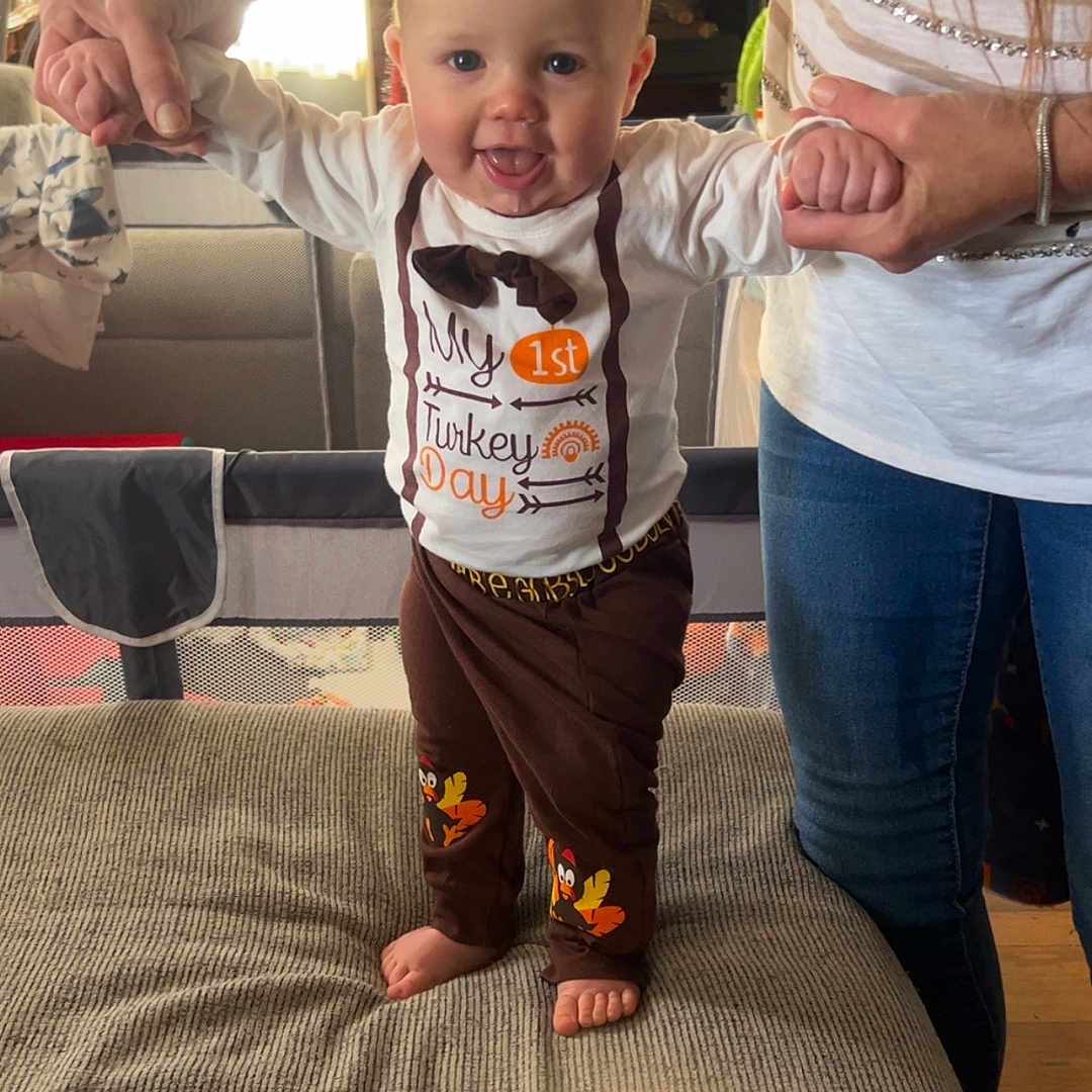 Damon is registered to the contest to win money with this photo: adult, baby, barefoot, child, clothing, couch, cute, footwear, happy, holding_hands, holiday, home, indoor, living_room, pants, person, shirt, smiling, standing, thanksgiving