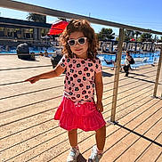 Kyla joined the competition — help win amazing prizes! child, girl, sunglasses, curly_hair, pink_skirt, patterned_shirt, sneakers, pool, outdoor, sunny, deck, railing, shadow, people, water, trees, umbrella, leisure, summer, fashion