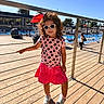 child, girl, sunglasses, curly_hair, pink_skirt, patterned_shirt, sneakers, pool, outdoor, sunny, deck, railing, shadow, people, water, trees, umbrella, leisure, summer, fashion
