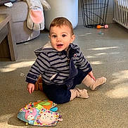 Nolan a rejoint le concours — aidez-le/la à gagner de superbes lots ! baby, carpet, casual_clothing, child, colorful_book, couch, floor, furniture, happy, indoor, plush_elephant, radiator, seated, shadow, smiling, socks, striped_hoodie, sunlight, toddler, toy