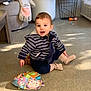 baby, carpet, casual_clothing, child, colorful_book, couch, floor, furniture, happy, indoor, plush_elephant, radiator, seated, shadow, smiling, socks, striped_hoodie, sunlight, toddler, toy