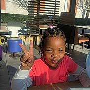 Juliana-Naomie participe au concours pour gagner de l'argent avec cette photo : child, peace_sign, indoor, table, sunlight, shadows, dining_area, chair, stool, pout, smile, hair_beads, window, blinds, casual, daylight, person, pink_shirt, long_sleeves, playful