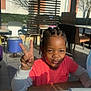 child, peace_sign, indoor, table, sunlight, shadows, dining_area, chair, stool, pout, smile, hair_beads, window, blinds, casual, daylight, person, pink_shirt, long_sleeves, playful