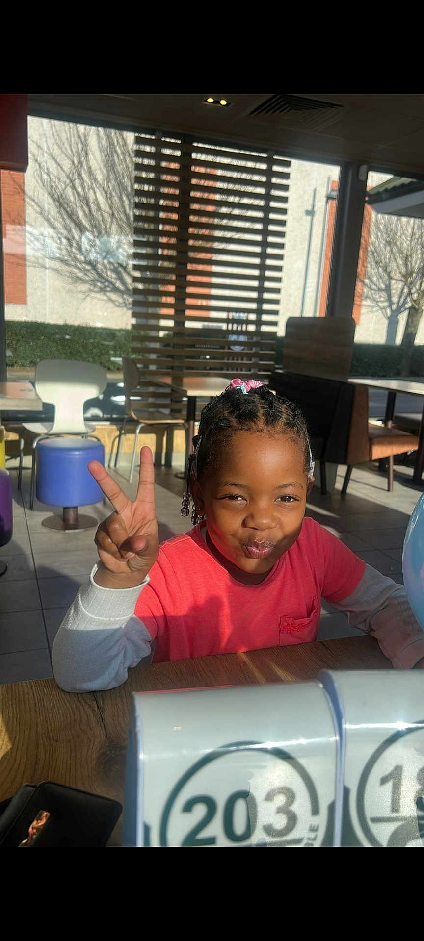 Juliana-Naomie participe au concours pour gagner de l'argent avec cette photo : child, peace_sign, indoor, table, sunlight, shadows, dining_area, chair, stool, pout, smile, hair_beads, window, blinds, casual, daylight, person, pink_shirt, long_sleeves, playful