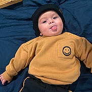 River is registered to the contest to win money with this photo: baby, child, beanie, black_hat, mustard_sweatshirt, smiley_face, tongue_out, lying_down, navy_blanket, playful, cute, infant, clothing, person, portrait, indoor, happy, face, headwear, casual