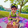 child, toddler, cake, birthday_cake, candles, number_candles, outdoor, park, tree, grass, blanket, headband, green_clothing, purple_clothing, playing, celebration, food, daylight, happy, cute