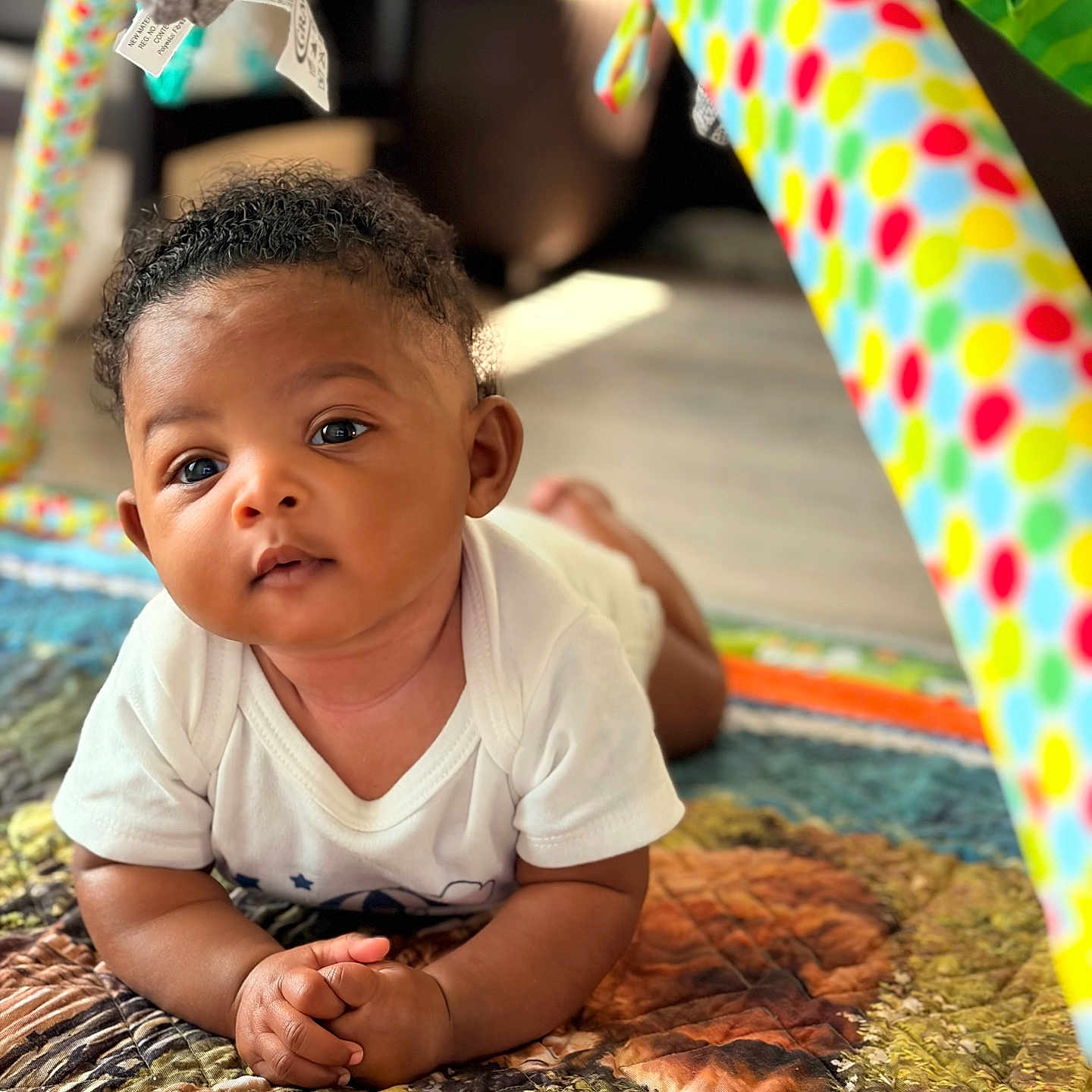 Dez joined the competition — help win amazing prizes! baby, infant, child, curly_hair, white_shirt, colorful_quilt, play_gym, toy, indoor, floor, hands, face, eyes, skin, crawling, tummy_time, portrait, cute, soft_light, homedecor
