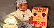 Dez is registered to the contest to win money with this photo: baby, infant, pajamas, cap, lamp, book, bed, gray_bedding, child, indoors, cute, sitting, face, expression, lighting, cozy, reading, storybook, soft_light, curious