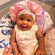 Samora is registered to the contest to win money with this photo: baby, child, smiling, pink_bonnet, onesie, blanket, hand, happy, infant, cute, indoors, portrait, person, skin, face, expression, seated, colorful, soft, cozy