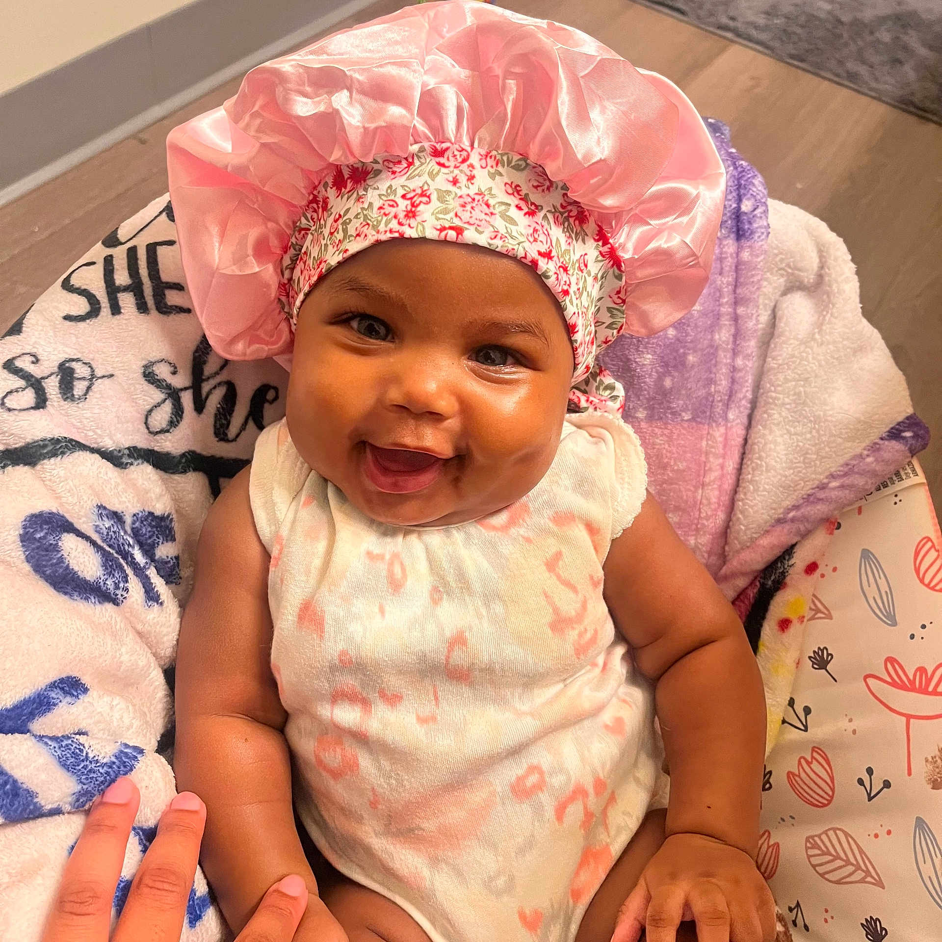 Samora is registered to the contest to win money with this photo: baby, child, smiling, pink_bonnet, onesie, blanket, hand, happy, infant, cute, indoors, portrait, person, skin, face, expression, seated, colorful, soft, cozy
