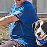 animal, blue_bandana, blue_shirt, building, casual_clothing, companion, daylight, dog, grass, happy, leisure, man, nature, outdoor, person, pet, smiling, sunny, tongue_out, tree