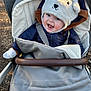 baby, child, stroller, hat, bear_hat, jacket, smile, happy, outdoor, cold_weather, mittens, blanket, seatbelt, face, person, infant, cute, cozy, daylight, nature