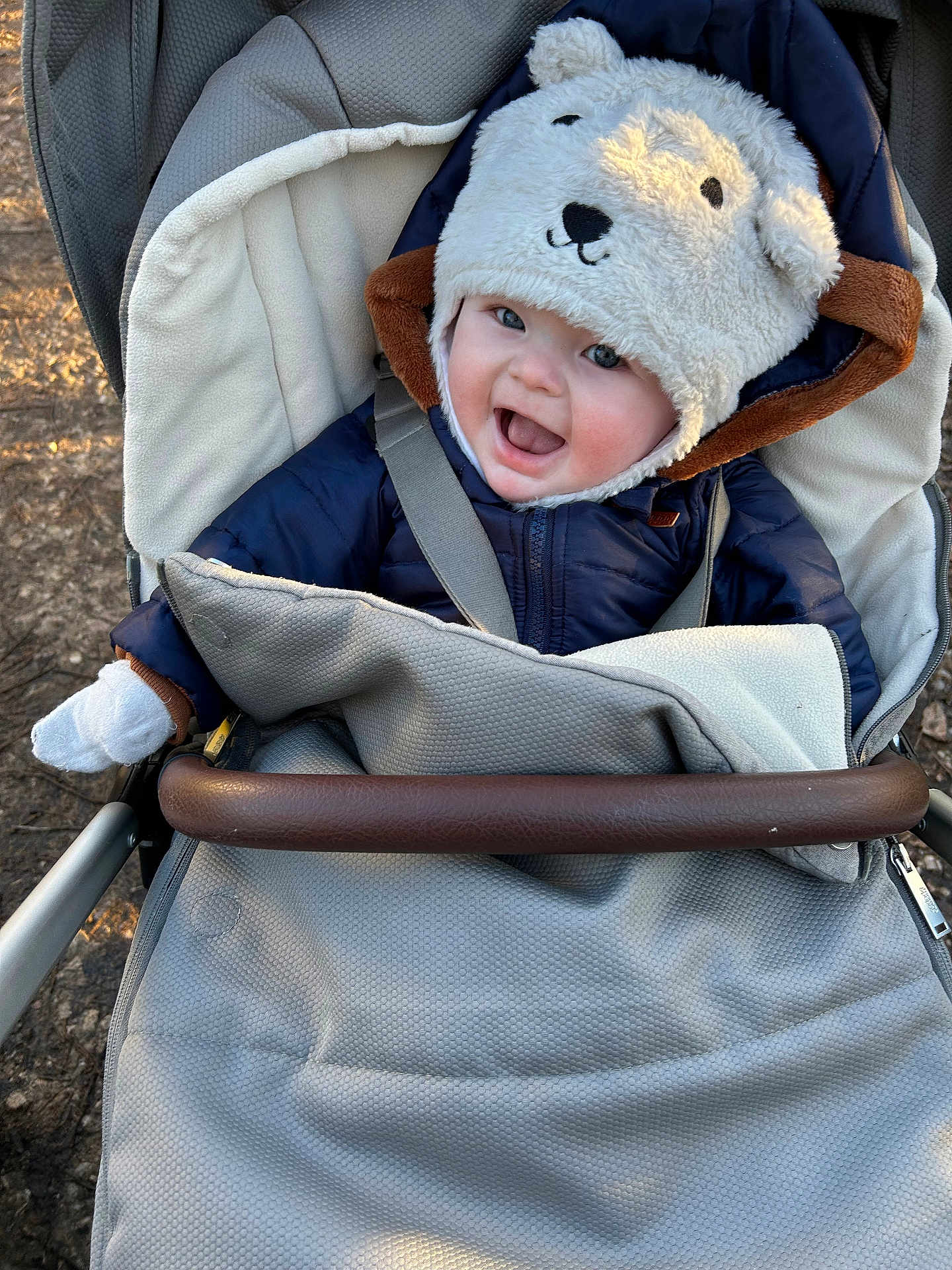 Teddy is registered to the contest to win money with this photo: baby, child, stroller, hat, bear_hat, jacket, smile, happy, outdoor, cold_weather, mittens, blanket, seatbelt, face, person, infant, cute, cozy, daylight, nature